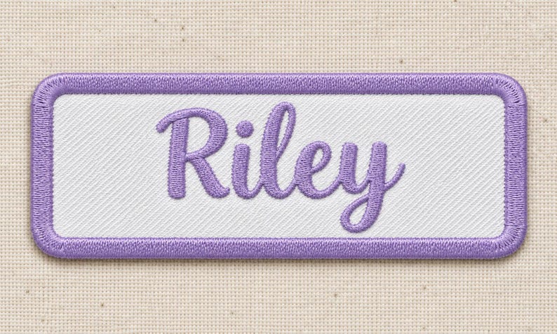 Close view of a complete white twill patch reading Riley in purple cursive embroidery with a purple satin-stitched border, lying on cream woven fabric. Fine diagonal fabric lines and individual thread stitches are visible.
