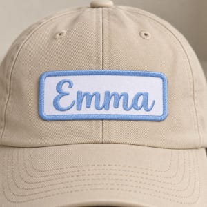 Close view of a beige baseball cap with a white rectangular name patch reading Emma in light blue cursive embroidery, surrounded by a matching blue stitched border.