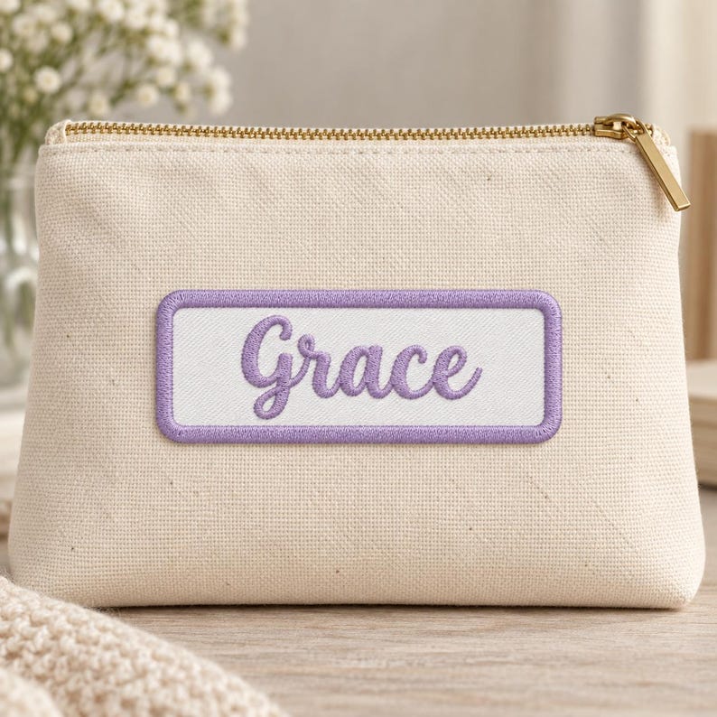 Cream canvas zipper pouch with a gold zipper and a white rectangular patch reading Grace in purple cursive embroidery, framed by a matching purple stitched border. Blurred white flowers appear behind it.