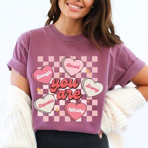 May include: A mauve t-shirt with a pink and white checkered background. The text "you are" is in a retro font with pink hearts surrounding it. Each heart has a positive affirmation written on it: "Loved", "Enough", "Strong", "Capable", and "Worthy".