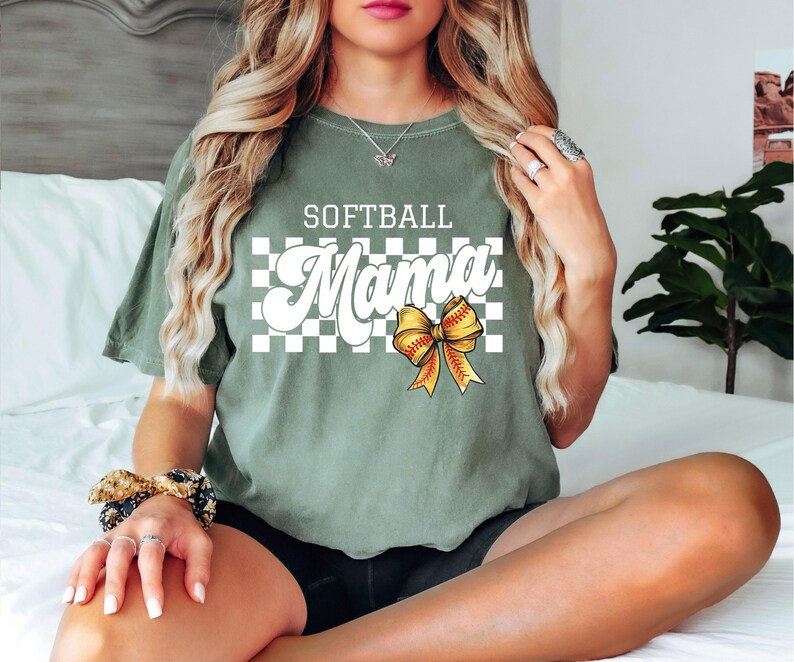 May include: A green t-shirt with the text "Softball Mama" in white letters with a checkered background and a yellow bow with a softball design.