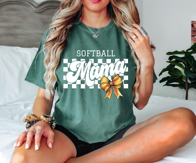 May include: A green t-shirt with a white checkered background and the text "Softball Mama" in a white retro font. A yellow and red softball bow is centered on the shirt.