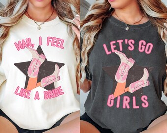 May include: Two white and gray t-shirts with pink text and graphics. The first shirt says "Man, I feel like a bride" with a graphic of a woman's legs wearing pink cowboy boots. The second shirt says "Let's go girls" with a graphic of a woman's legs wearing pink cowboy boots.