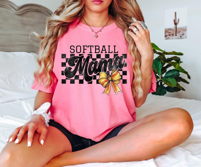 May include: Pink softball t-shirt with black and white checkered pattern and the text "Softball Mama" in black with a yellow and black softball bow.