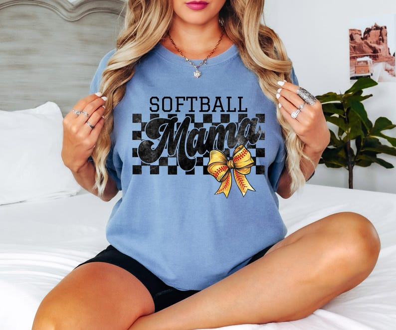 May include: A light blue t-shirt with a black and white checkered pattern and the text "Softball Mama" in black. A yellow and red softball bow is centered on the checkered pattern.