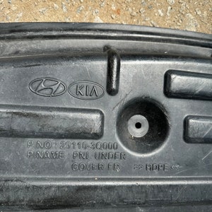 Puede incluir: Pieza de automóvil de plástico negro con los logotipos de Hyundai y Kia. El número de pieza es 29110-3Q000, y el nombre es PNL UNDER COVER FR. El material es HDPE.