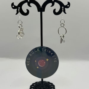 May include: A pair of silver earrings on a black earring stand. The earrings are both dangling and feature a charm. One earring has a charm of a handcuff and the other earring has a charm of a set of keys. The stand has a black and white sticker that says "Red Rock Trinkets" with a red moon and stars.
