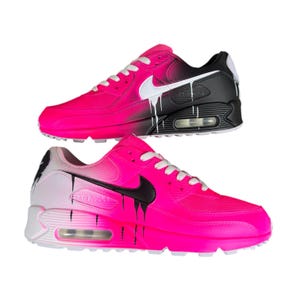 May include: Two pairs of Nike Air Max 90 sneakers. The shoes are primarily hot pink with white laces and a white Nike swoosh. One shoe has a black heel and the other has a white heel. Both shoes have a drip paint design.