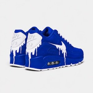 May include: A pair of royal blue sneakers featuring a white Nike Air logo on the heel and a white drip pattern design extending from the heel and side swoosh. The shoes include white laces, a thick blue midsole, and a visible air cushioning unit.