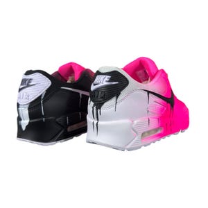 May include: Pair of Nike Air Max 90 sneakers. One shoe is black with white accents and dripping paint effect. The other shoe is white and fades to pink, also with a dripping paint effect. Both shoes have a pink interior.