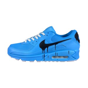 May include: A light blue Nike Air Max 90 sneaker with a black Nike swoosh and black paint drip design. The shoe has white laces and a white sole. The Air Max logo is visible on the side.