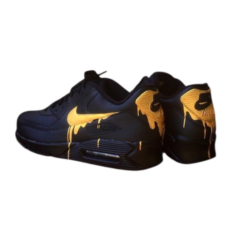 May include: Black Nike Air Max 90 sneakers with gold accents. The iconic Nike swoosh and heel detail are gold, with a dripping paint effect. The shoes have a black sole and laces, and the Air Max logo is visible.