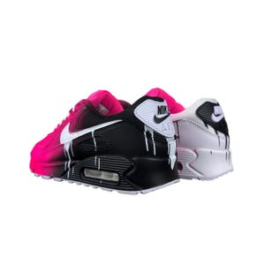 May include: Pair of Nike Air Max 90 sneakers. One shoe is black and pink with white accents and dripping paint effect. The other shoe is white and pink with black accents and dripping paint effect. The Nike logo is visible on both shoes.