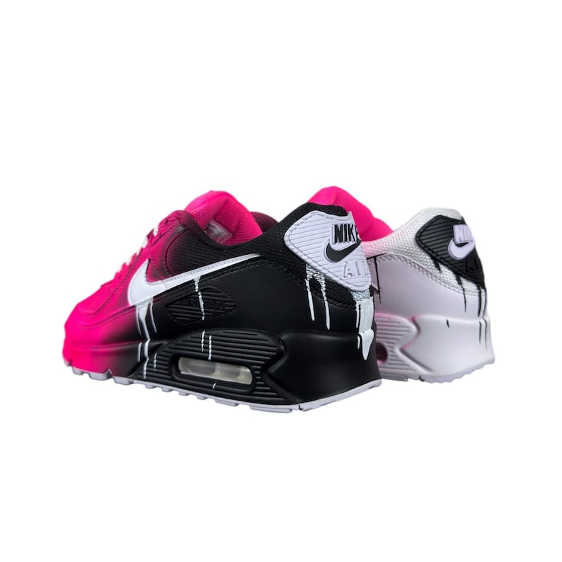 May include: Pair of Nike Air Max 90 sneakers. One shoe is black and pink with white accents and dripping paint effect. The other shoe is white and pink with black accents and dripping paint effect. The Nike logo is visible on both shoes.