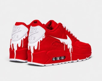 May include: A pair of bright red sneakers featuring a white Nike Air logo on the heel and a white swoosh on the side. The shoes are decorated with a white paint drip effect that extends from the heel and swoosh down onto the red midsole.