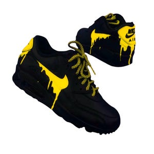 Puede incluir: Zapatillas Nike Air Max 90 negras y amarillas. Las zapatillas tienen una base negra con detalles amarillos, incluyendo el logo de Nike y un dise&ntilde;o de pintura goteando. Los cordones son amarillos con motas negras.
