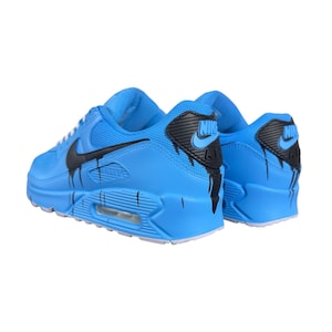 May include: A pair of light blue Nike Air Max 90 sneakers with black accents. The Nike swoosh and heel detail are black, with black paint drip design. The shoes have white soles.