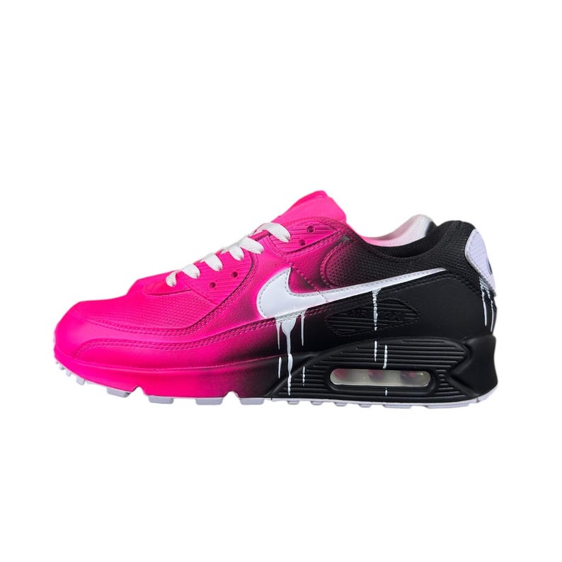 May include: A pink and black Nike Air Max 90 sneaker. The shoe transitions from bright pink to black, with white accents including the Nike swoosh and dripping paint effect. White laces and a white sole complete the design.