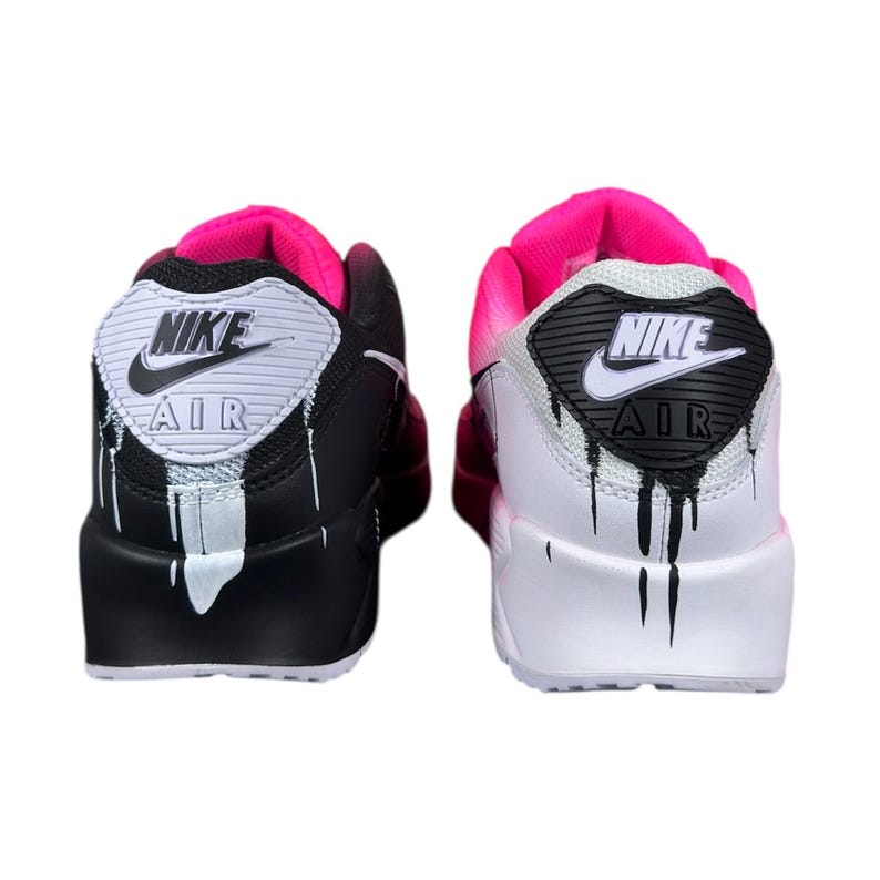 May include: Pair of Nike Air Max 90 sneakers. One shoe is black with white accents and dripping paint effect. The other is white with black accents and a dripping paint effect. Both have pink and white details.