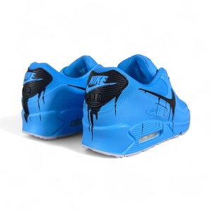 May include: A pair of light blue athletic shoes with black accents. The shoes feature the Nike logo and a unique dripping paint design. The soles are white, and the overall style is sporty and modern.