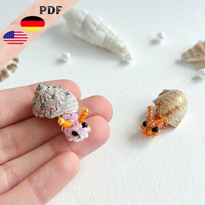Könnte beinhalten: Zwei Perlen-Einsiedlerkrebse, einer rosa und einer orange, sitzen in ihren Muscheln. Die Muscheln sind braun und weiß mit einem gefleckten Muster. Die Krebse sind aus kleinen Perlen gefertigt und haben schwarze Augen.
