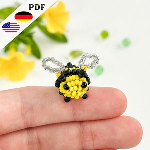 Può includere: Una piccola ape di perline con perline nere e gialle e ali argentate. L'ape è seduta su un dito.