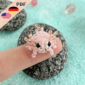 Könnte beinhalten: Ein rosa Perlen-Axolotl-Anhänger mit schwarzen Augen. Der Anhänger ist aus kleinen, runden Perlen gefertigt und etwa so groß wie ein Fingernagel.