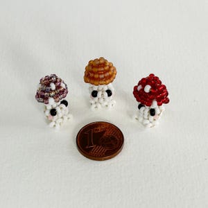 May include: Three small beaded mushroom figurines with white bodies, black eyes, and pink cheeks. The mushrooms have different colored tops: purple, brown, and red.
