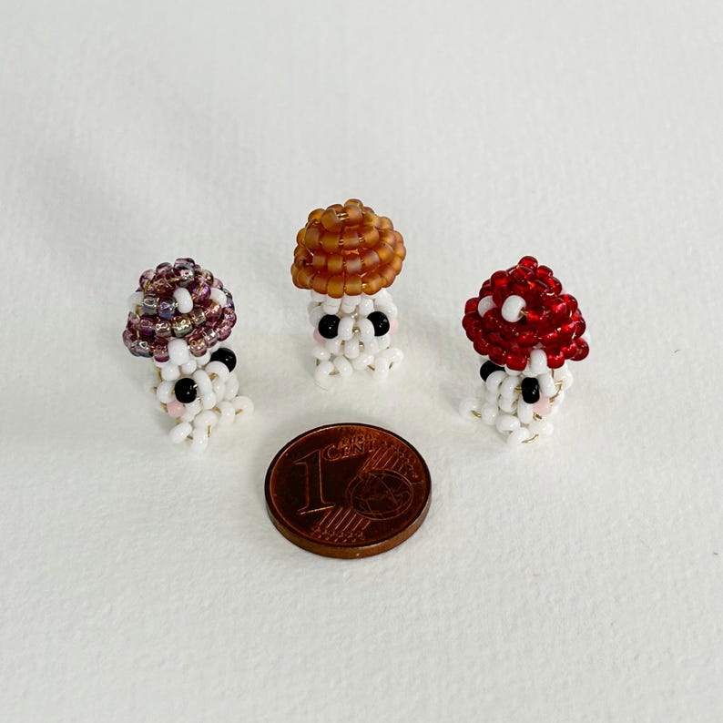 May include: Three small beaded mushroom figurines with white bodies, black eyes, and pink cheeks. The mushrooms have different colored tops: purple, brown, and red.