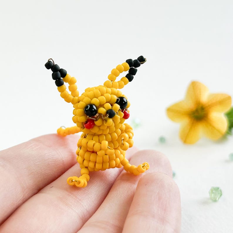 May include: A yellow beaded Pikachu figure with black-tipped ears, black eyes, and a red mouth. The figure is posed with its arms outstretched and legs extended. The figure is held in a hand, with a yellow flower and green beads in the background.