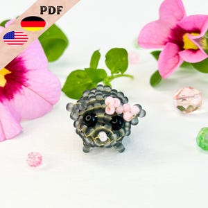 Puede incluir: Un pequeño animal gris de cuentas con ojos negros y una corona de flores rosas. El animal está sentado sobre una superficie blanca.