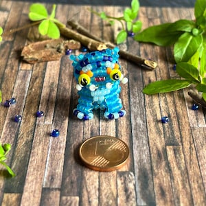 May include: A handmade blue beaded creature with yellow and white details, set on a wooden surface. The creature features large yellow eyes and a small red mouth. A coin is positioned nearby.