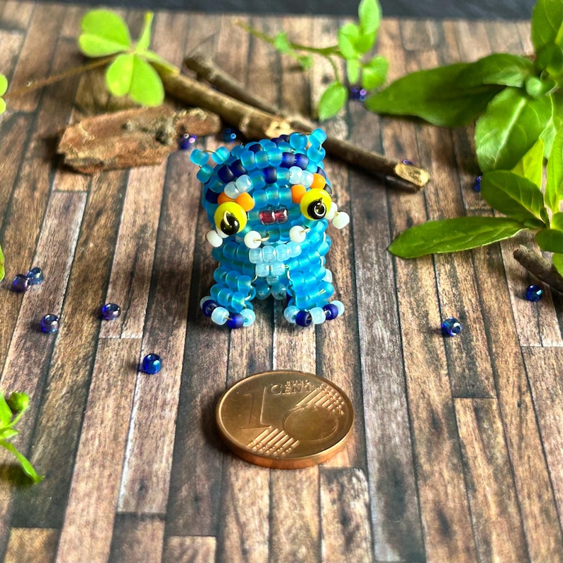 May include: A handmade blue beaded creature with yellow and white details, set on a wooden surface. The creature features large yellow eyes and a small red mouth. A coin is positioned nearby.