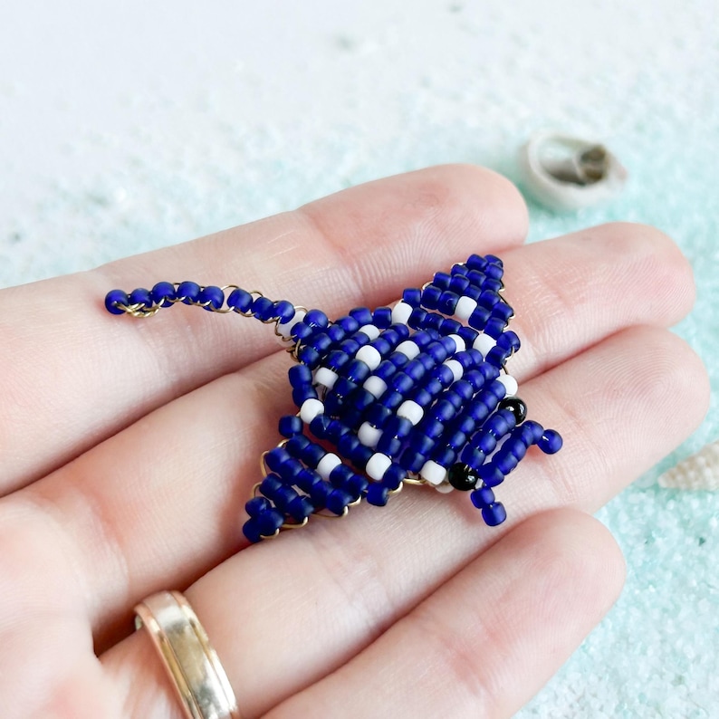May include: A handmade beaded stingray pin. The pin features a blue, white, and black beaded design with a gold-colored wire frame. The stingray is held in a hand, showcasing the intricate beadwork and craftsmanship.