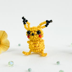 May include: A handcrafted yellow beaded Pikachu figure with black ears and eyes, and red cheeks. The figure is made of small, round beads and has a detailed design. The figure is sitting on a white surface.