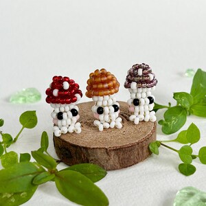 May include: Three miniature mushroom figurines made of white beads with different colored tops. The mushrooms are sitting on a small wooden slice. The mushrooms have black bead eyes and pink bead cheeks.