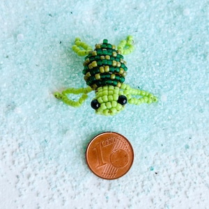 May include: A handmade beaded turtle in shades of green and dark green, with black bead eyes. The turtle is placed on a light blue surface with a 1 Euro cent coin for size comparison. The turtle's body is made of small beads.