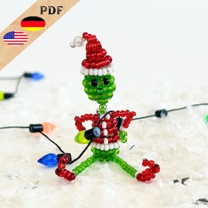 Puede incluir: Una figura de Grinch verde hecha de cuentas que lleva un gorro de Papá Noel rojo y blanco y sostiene una cadena de luces navideñas. La figura está sentada sobre una superficie blanca.