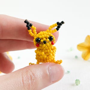 May include: A handmade yellow beaded Pikachu figure with black ears, black eyes, and red cheeks. The small figure is held between two fingers. The figure is made of small yellow beads, with black beads for the ears and eyes.