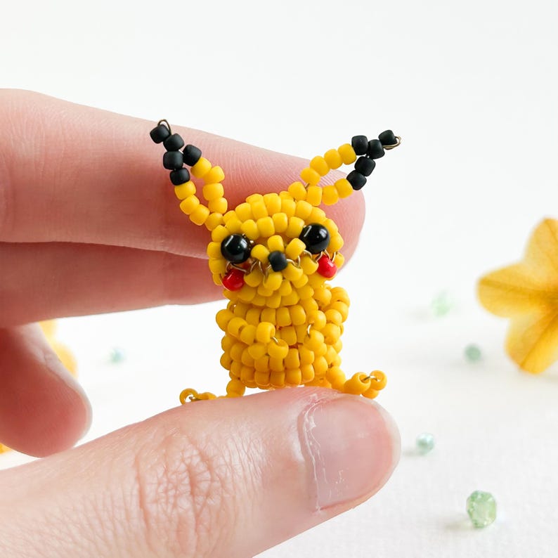 May include: A handmade yellow beaded Pikachu figure with black ears, black eyes, and red cheeks. The small figure is held between two fingers. The figure is made of small yellow beads, with black beads for the ears and eyes.
