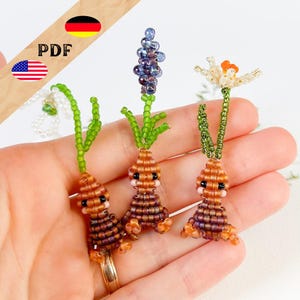 May include: Three beaded flower and vegetable charms. The charms include a carrot, a purple flower, and a white flower with an orange center. The carrot has a brown body, green stem, and black eyes. The charms are held in a hand.