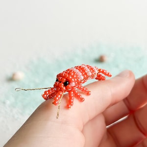 May include: A handcrafted beaded shrimp figurine. The shrimp is primarily red with white stripes and black eyes. It has delicate wire antennae and legs, and is made of small, round beads. The figurine is a miniature representation of a shrimp.