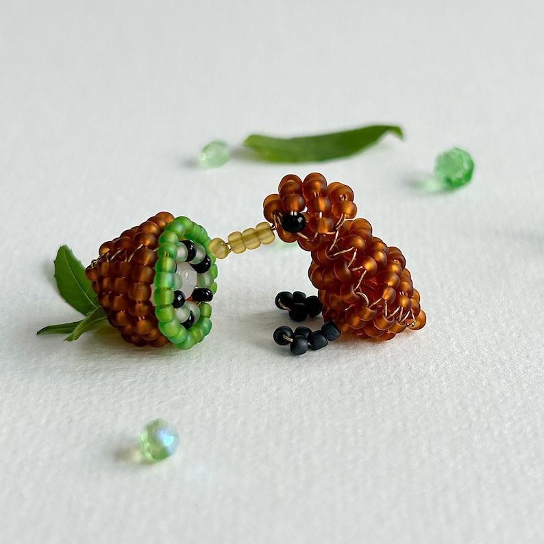 K&ouml;nnte beinhalten: Eine braune und gr&uuml;ne Perlenfigur eines Kiwi-Vogels, der eine Perlen-Kiwi-Frucht frisst. Der Kiwi-Vogel ist braun mit schwarzen Perlenaugen und die Kiwi-Frucht ist gr&uuml;n mit einer wei&szlig;en Mitte.