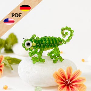 May include: A small, handcrafted chameleon figurine constructed from vibrant green glass beads and thin gold-toned wire. The creature features a curled tail, four legs, and two prominent white eyes with black pupils, standing on a smooth white stone.