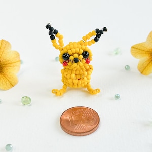 May include: A handmade yellow beaded Pikachu figure with black and red accents. The figure is posed with its arms and legs extended. A penny is next to the figure for size comparison. The background is white with scattered green beads and yellow flowers.