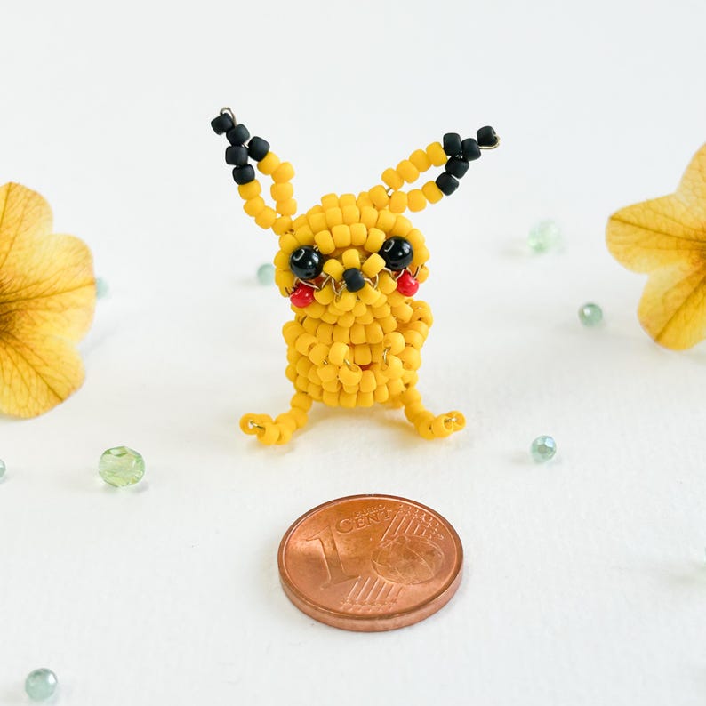 May include: A handmade yellow beaded Pikachu figure with black and red accents. The figure is posed with its arms and legs extended. A penny is next to the figure for size comparison. The background is white with scattered green beads and yellow flowers.
