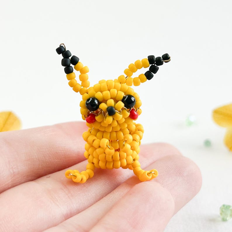 May include: A handmade Pikachu figurine crafted from yellow beads, with black bead accents on the ears and eyes, and red cheeks. The small, detailed figure is posed sitting down, showcasing the character's iconic features.
