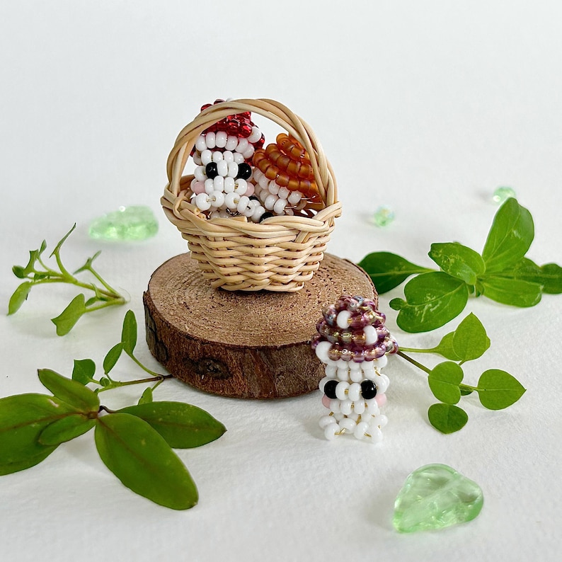 May include: Three beaded mushroom figurines in a miniature wicker basket. The mushrooms are white with red, orange, and purple accents. One mushroom is sitting outside the basket.