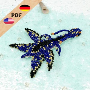 May include: A beaded starfish in blue, black, and gold. The handcrafted sea creature is detailed with small beads and has a textured appearance. The starfish is on a light blue surface, with a seashell in the background.
