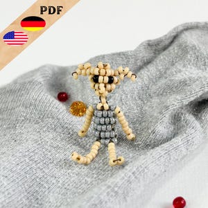 Könnte beinhalten: Eine kleine Perlenfigur eines Wesens mit großen Ohren, einem grauen Oberkörper und beigefarbenen Gliedmaßen. Die Figur sitzt auf einer grauen Stoffoberfläche.